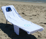 A white Ostrich custom fitted towel with a pocket, fitted on an Ostrich chaise lounge on a sandy beach.