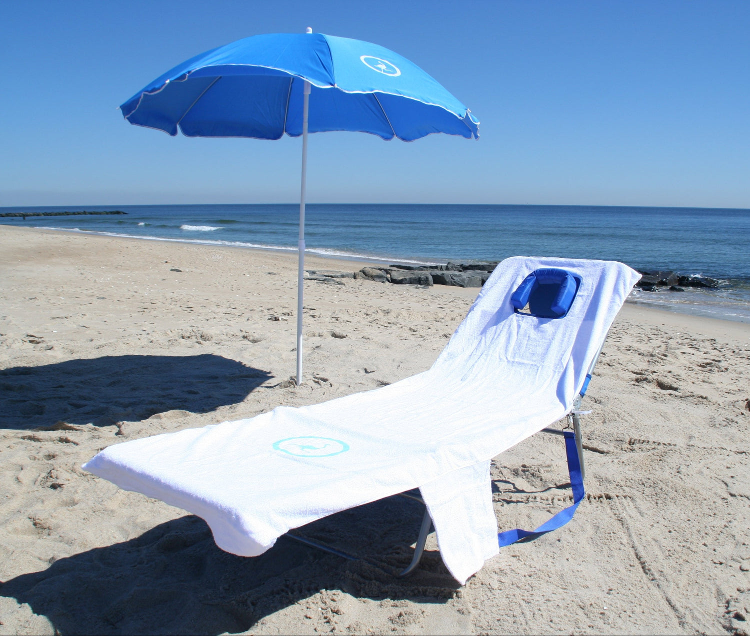 Ostrich chaise lounge with a custom fitted towel, in front of a blue Ostrich umbrella, on a sandy beach with ocean in the background